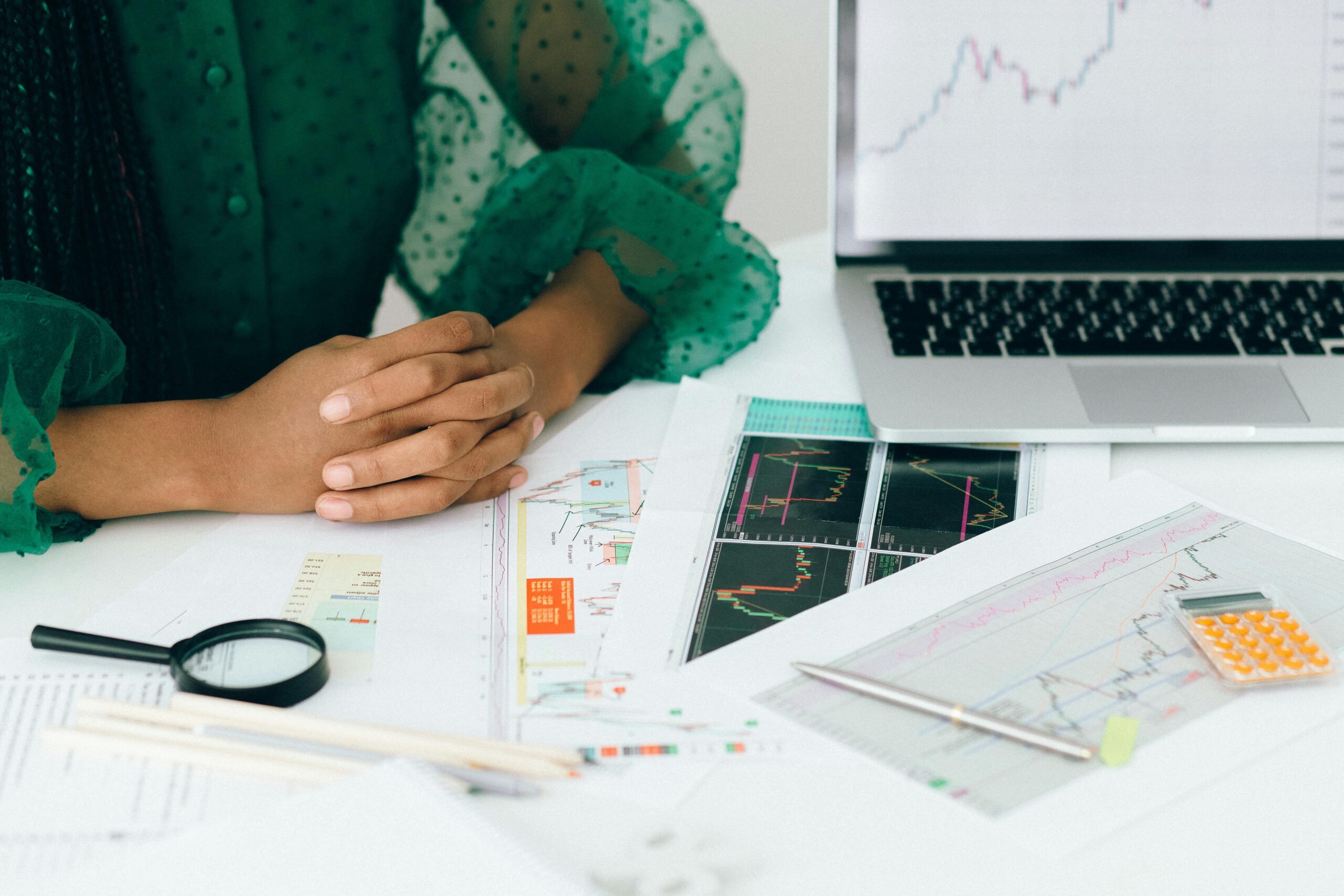 Person in grüner Bluse sitzt an einem Schreibtisch mit Finanzdiagrammen, Laptop, Taschenrechner und Lupe